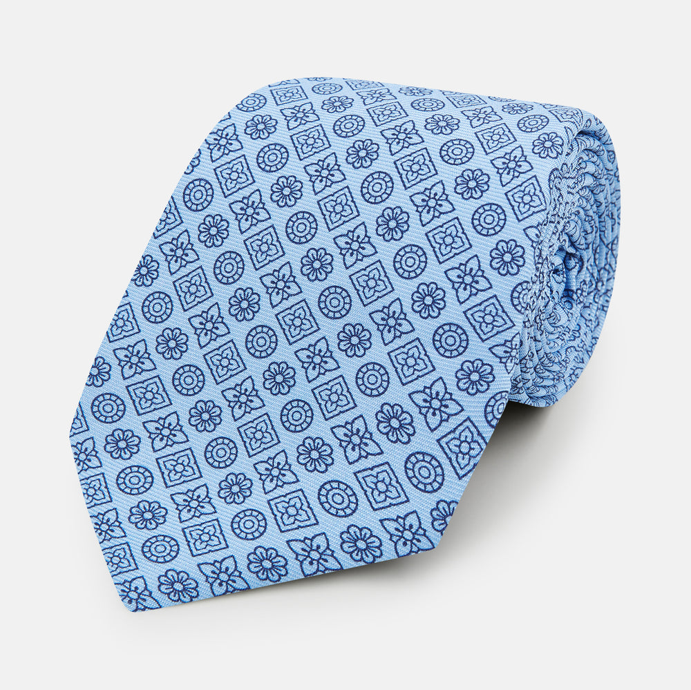 The Turnbull & Asser Blue Floral Tile Pattern Printed Silk Tie features a light blue geometric and floral design in dark blue, shown rolled up on a white background.