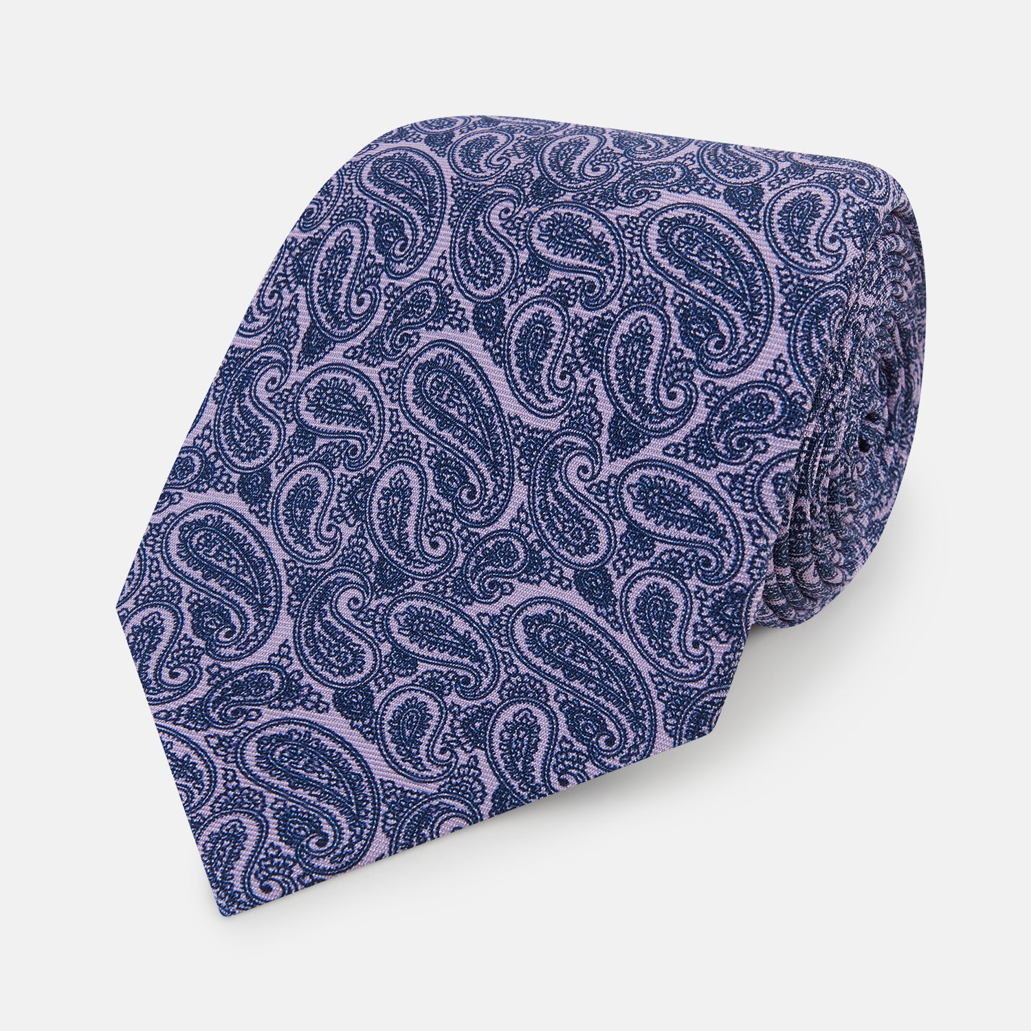 View 1 of product The Turnbull & Asser Lilac Paisley Silk Tie is shown rolled up, featuring a blue paisley pattern on a light purple background and displayed on a white surface.