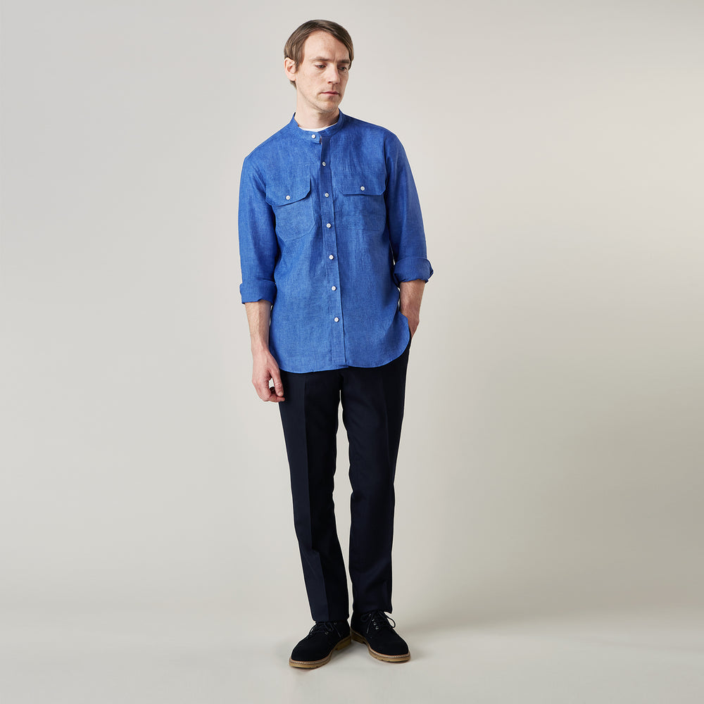 A man faces forward with a neutral expression, hands in his pockets, wearing the Turnbull & Asser Plain Indigo Linen Weekend Fit Shirt with a stand collar, styled with dark pants and black shoes against a plain light background.