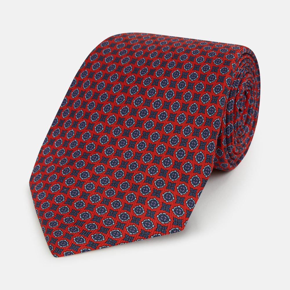 The Turnbull & Asser Red and Blue Geometric Floral Silk Tie features a red background with small blue and white circular floral patterns, exemplifying British craftsmanship.
