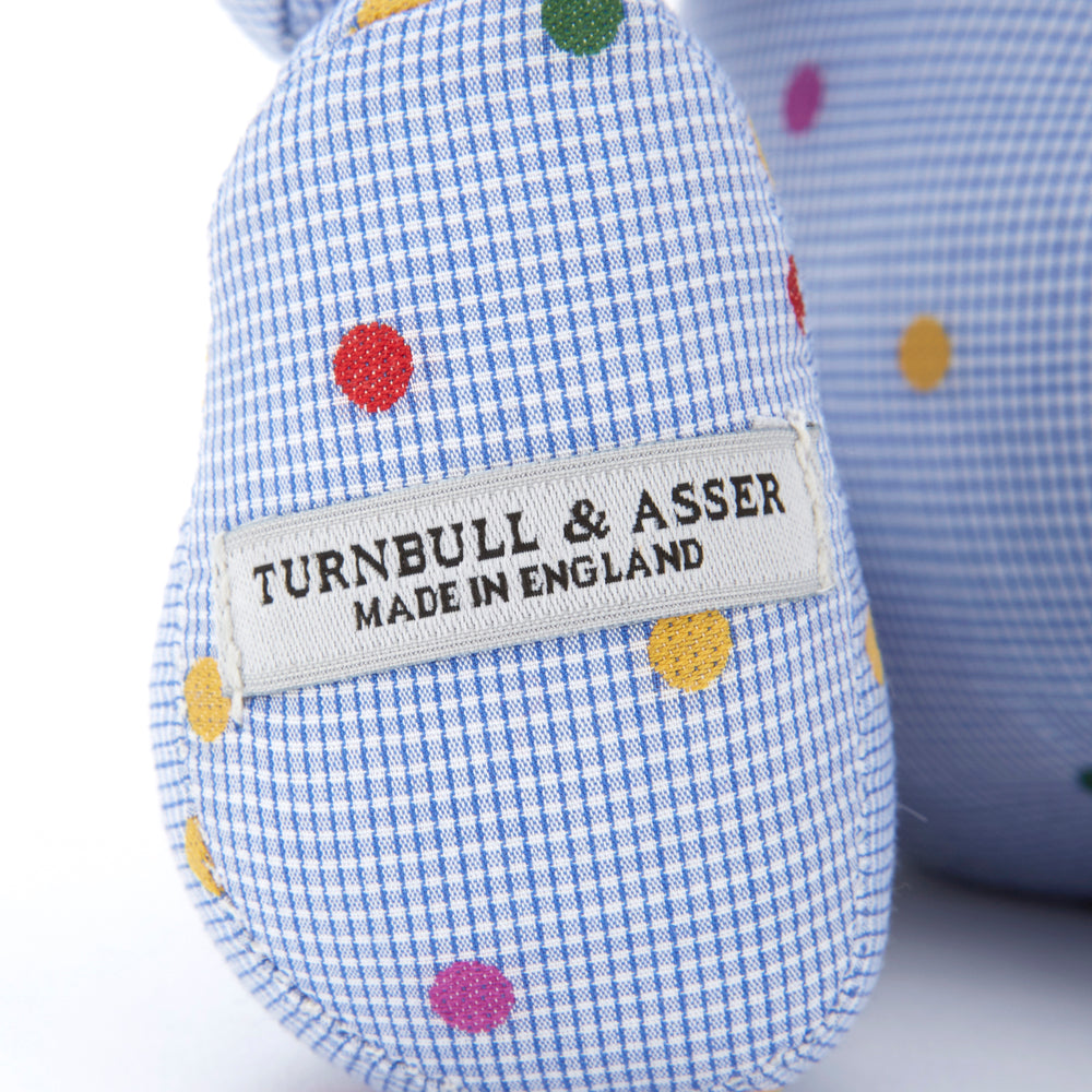 The Turnbuline Cotton Spot Teddy Bear by Turnbull & Asser is a blue gingham bear with colorful polka dots, black button eyes, a black nose, and a white leg label, shown sitting upright in profile facing left.