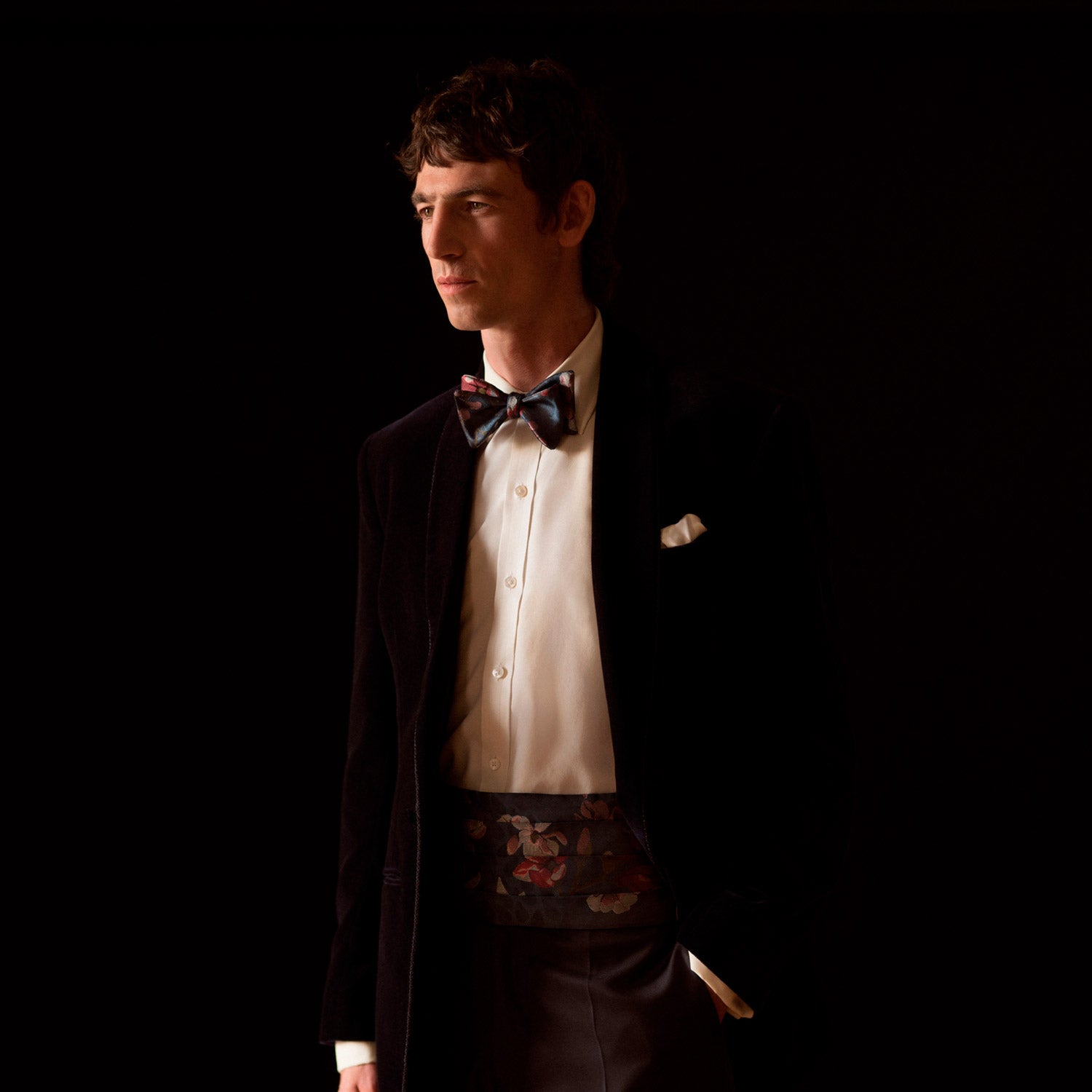 View 1 of product A man wearing a Turnbull & Asser Exclusive Silk Magnolia Cummerbund, paired with a black tuxedo jacket, white dress shirt, and patterned bow tie, stands against a dark background with a serious look.