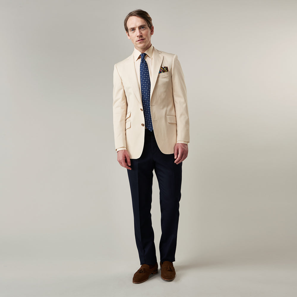 A man in a Turnbull & Asser Beige Cotton Barrington Blazer, light yellow shirt, blue patterned tie, and dark trousers stands against a plain light background, with a pocket square visible and a neutral expression.