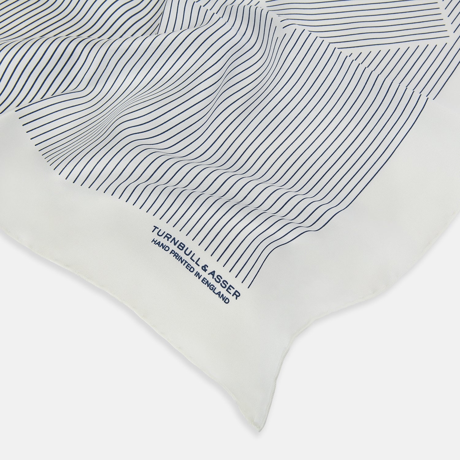 View 2 of product The White Stripe Silk Pocket Square by Turnbull & Asser features vertical navy pinstripes and “TURNBULL & ASSER HAND PRINTED IN ENGLAND” in blue at the lower right, with one corner neatly folded for a classic British look.