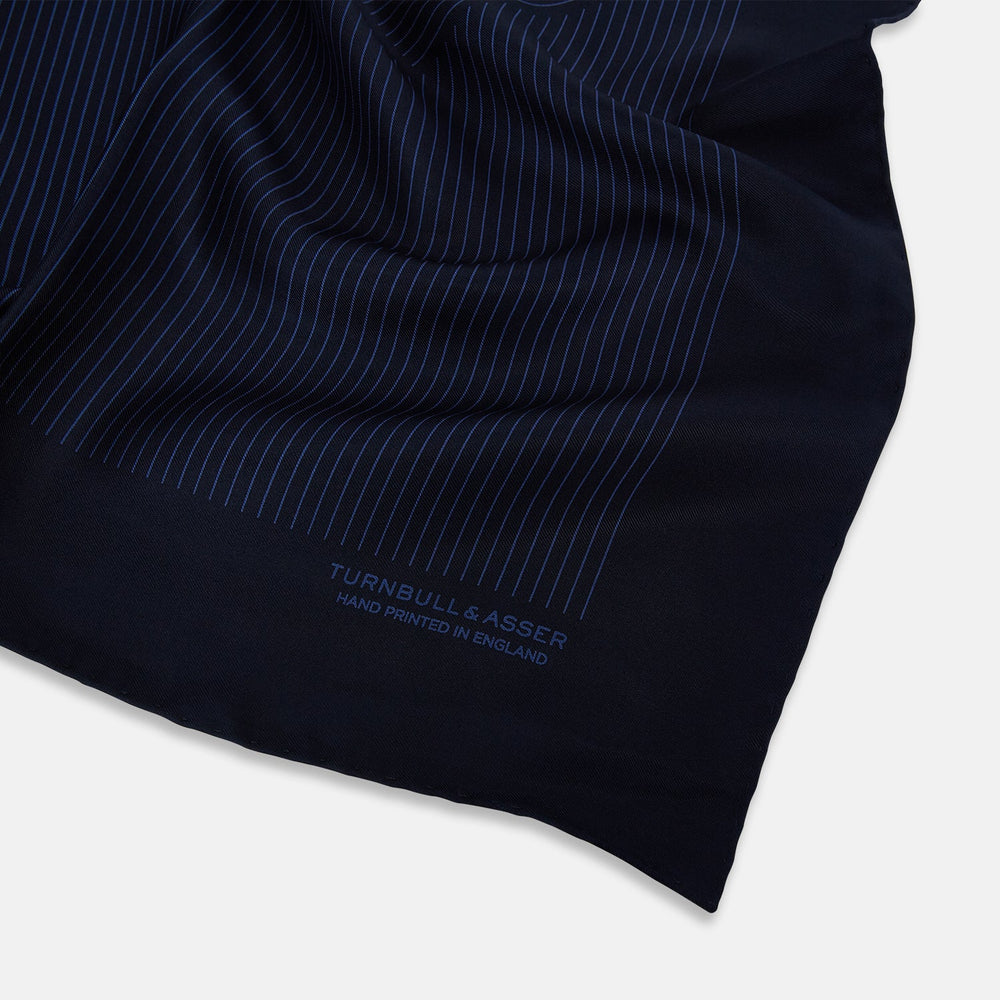 The Navy Stripe Silk Pocket Square by Turnbull & Asser features a dark navy silk with thin light blue stripes and hand-rolled edges, folded to reveal both sides and “TURNBULL & ASSER HAND PRINTED IN ENGLAND” in the corner.