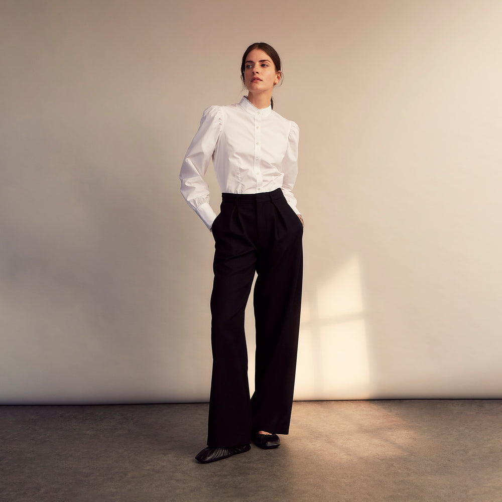 A woman stands in profile, looking down with hands in her pockets, wearing the Turnbull & Asser Turnbull and The Deck White Cotton Expressionist Shirt, paired with high-waisted black-and-white houndstooth pants.
