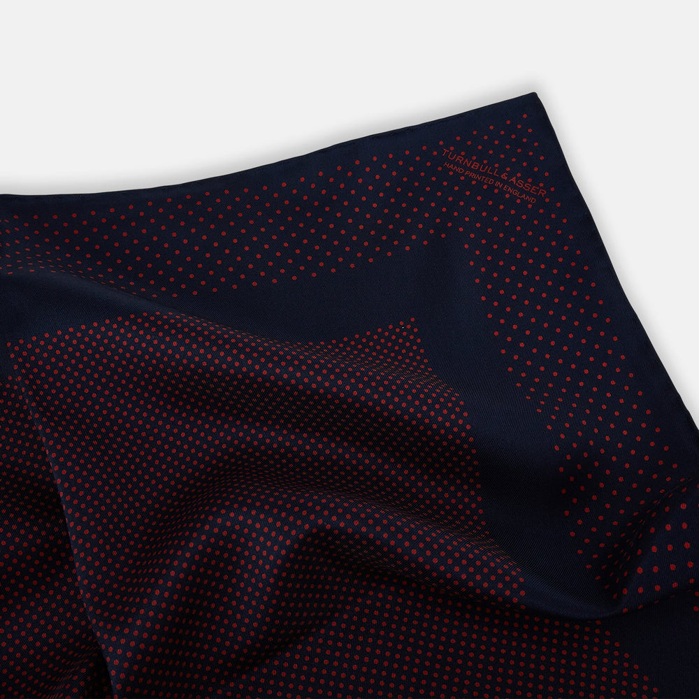 The Turnbull & Asser Navy and Red Dot Silk Pocket Square features a dark navy blue base with small red polka dots on the border and inner pattern, all handmade and neatly folded.