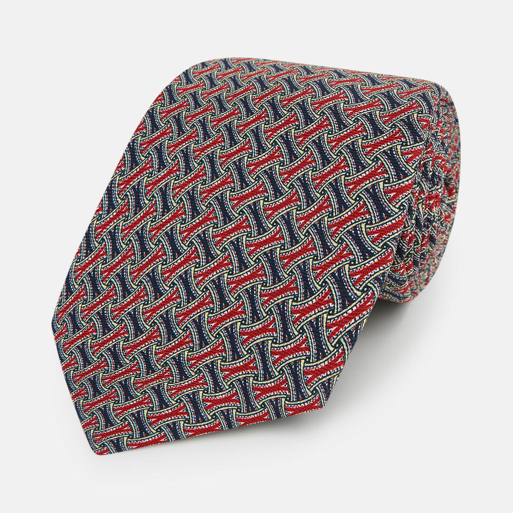 The Turnbull & Asser Red and Navy Weave-Patterned Printed Silk Tie features an intricate geometric design in red, blue, and white. Displayed rolled-up against a plain white background, this classic tie is crafted from luxurious silk.