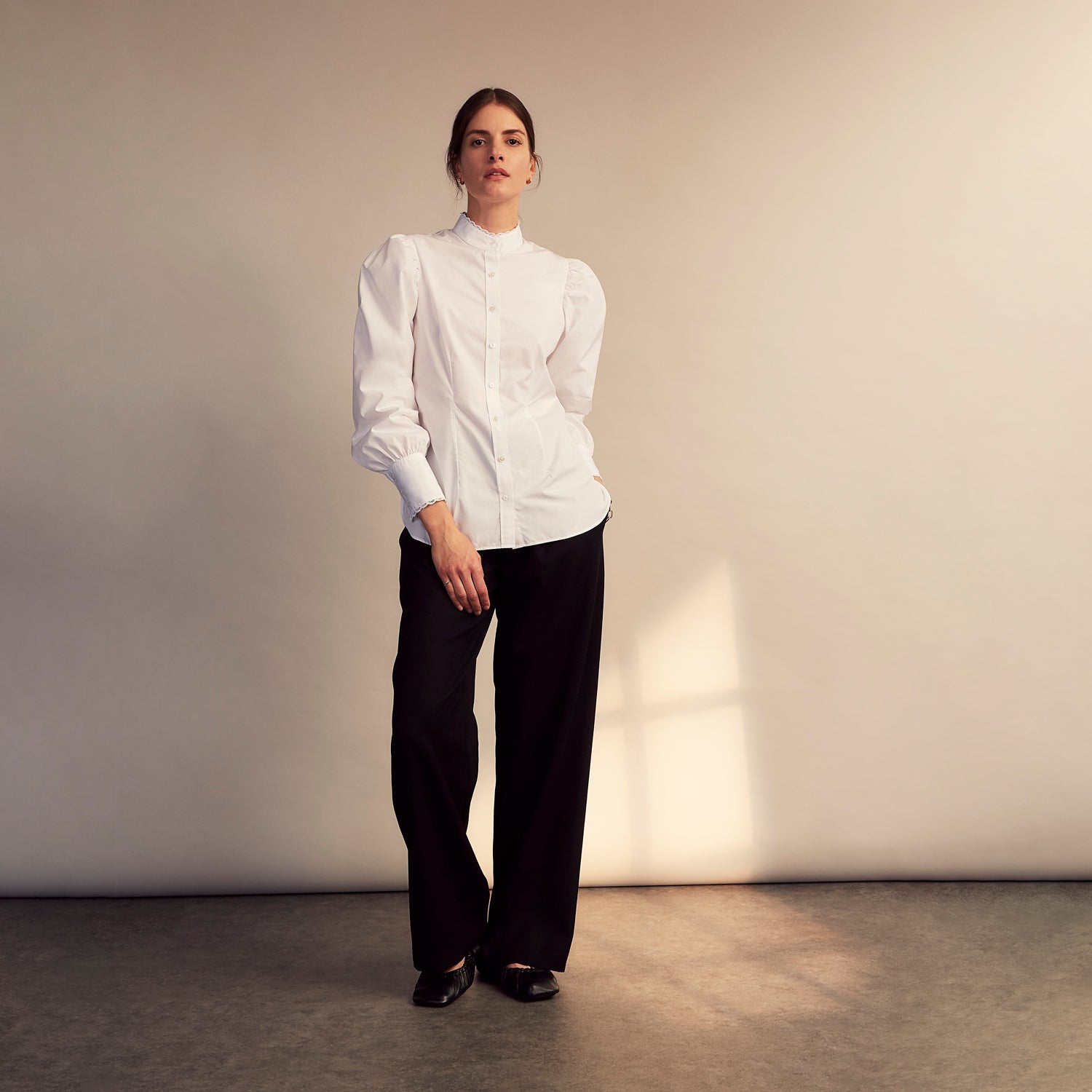 View 4 of product A woman stands in profile, looking down with hands in her pockets, wearing the Turnbull & Asser Turnbull and The Deck White Cotton Expressionist Shirt, paired with high-waisted black-and-white houndstooth pants.