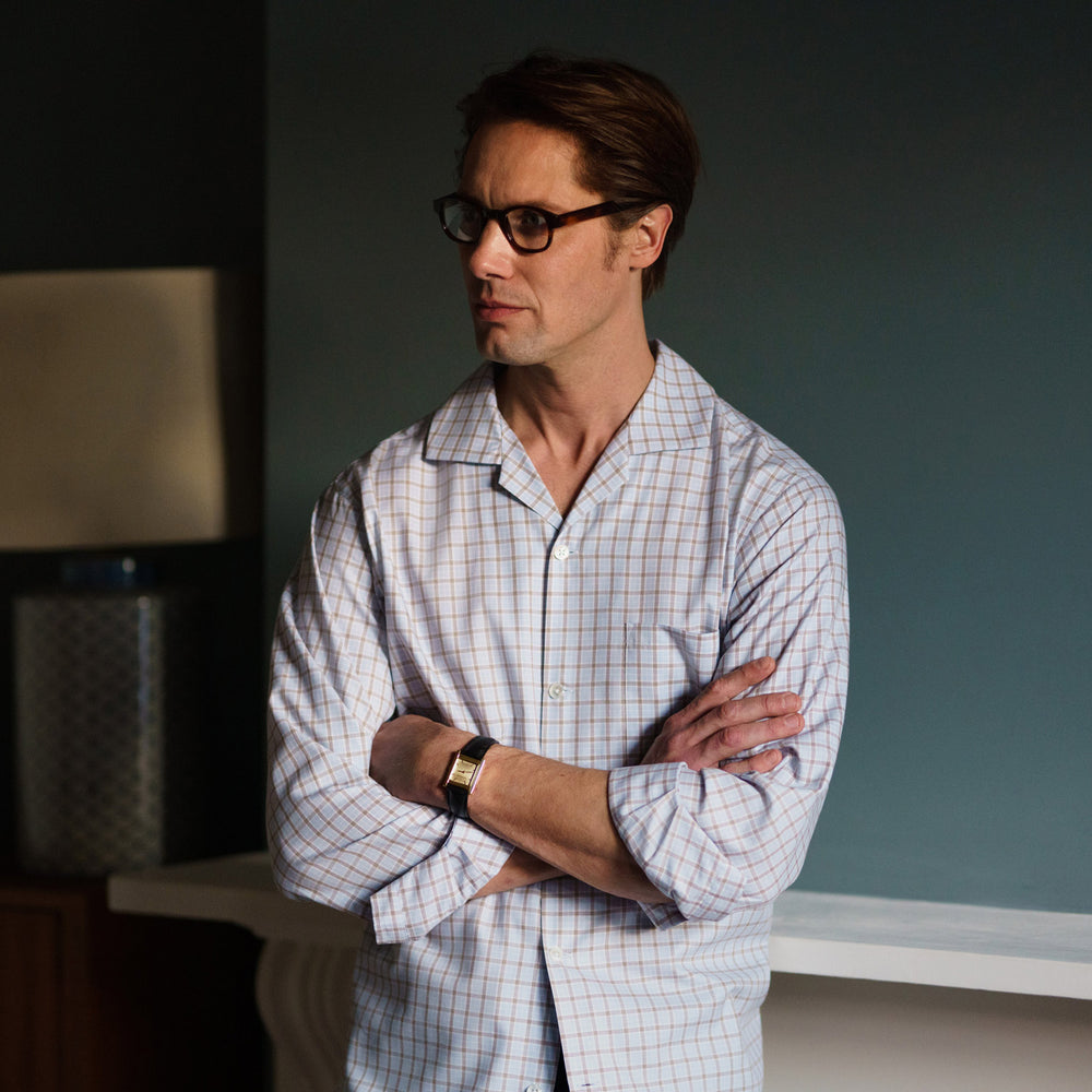 A man wearing glasses and a Pale Blue Multi Check Cashmerello Holiday Fit Winnington Shirt by Turnbull & Asser leans casually against a white countertop, looking at the camera with a neutral expression.
