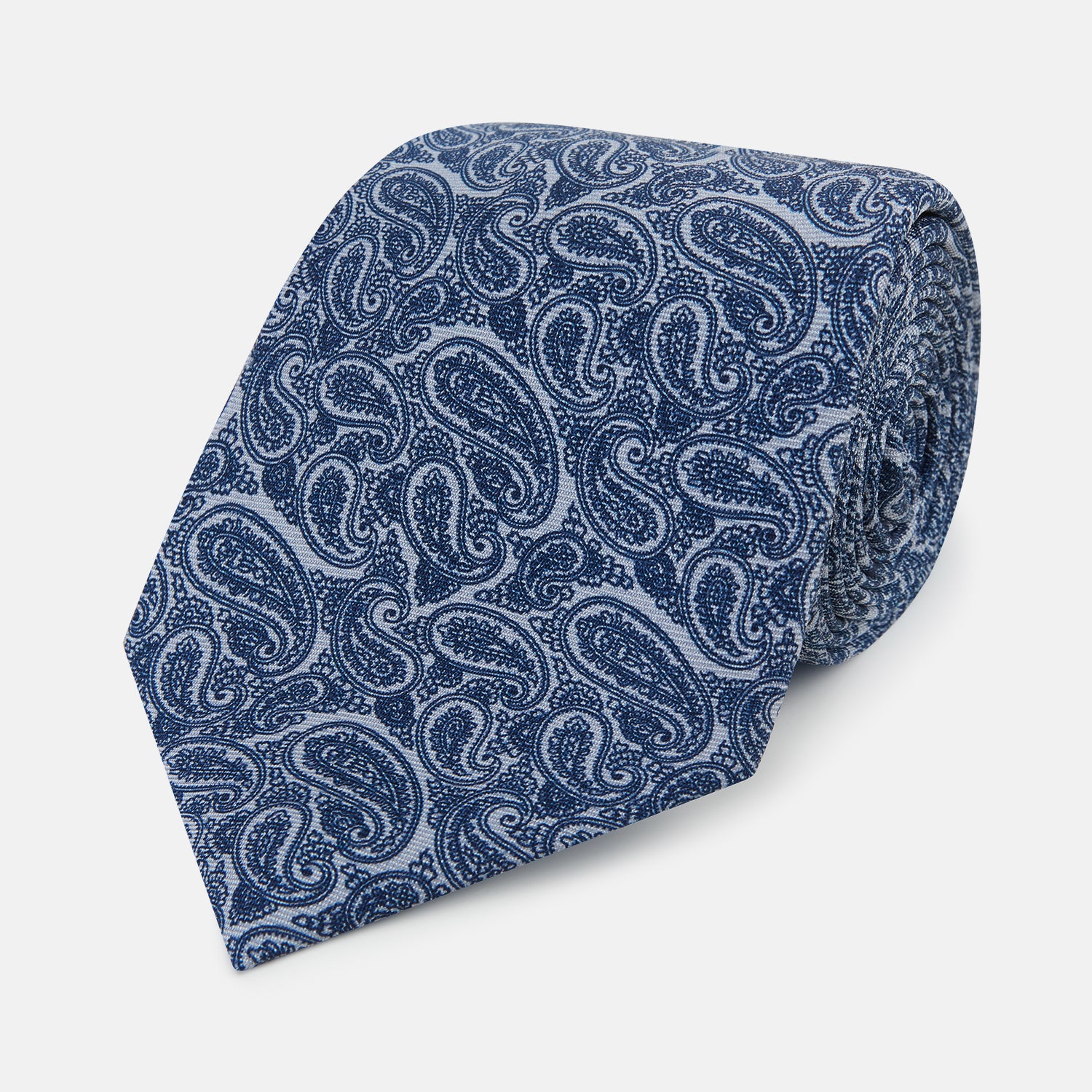 View 1 of product The Turnbull & Asser Pale Blue Paisley Printed Silk Tie is displayed rolled up on a white background, showcasing its intricate paisley pattern in varied blue tones.