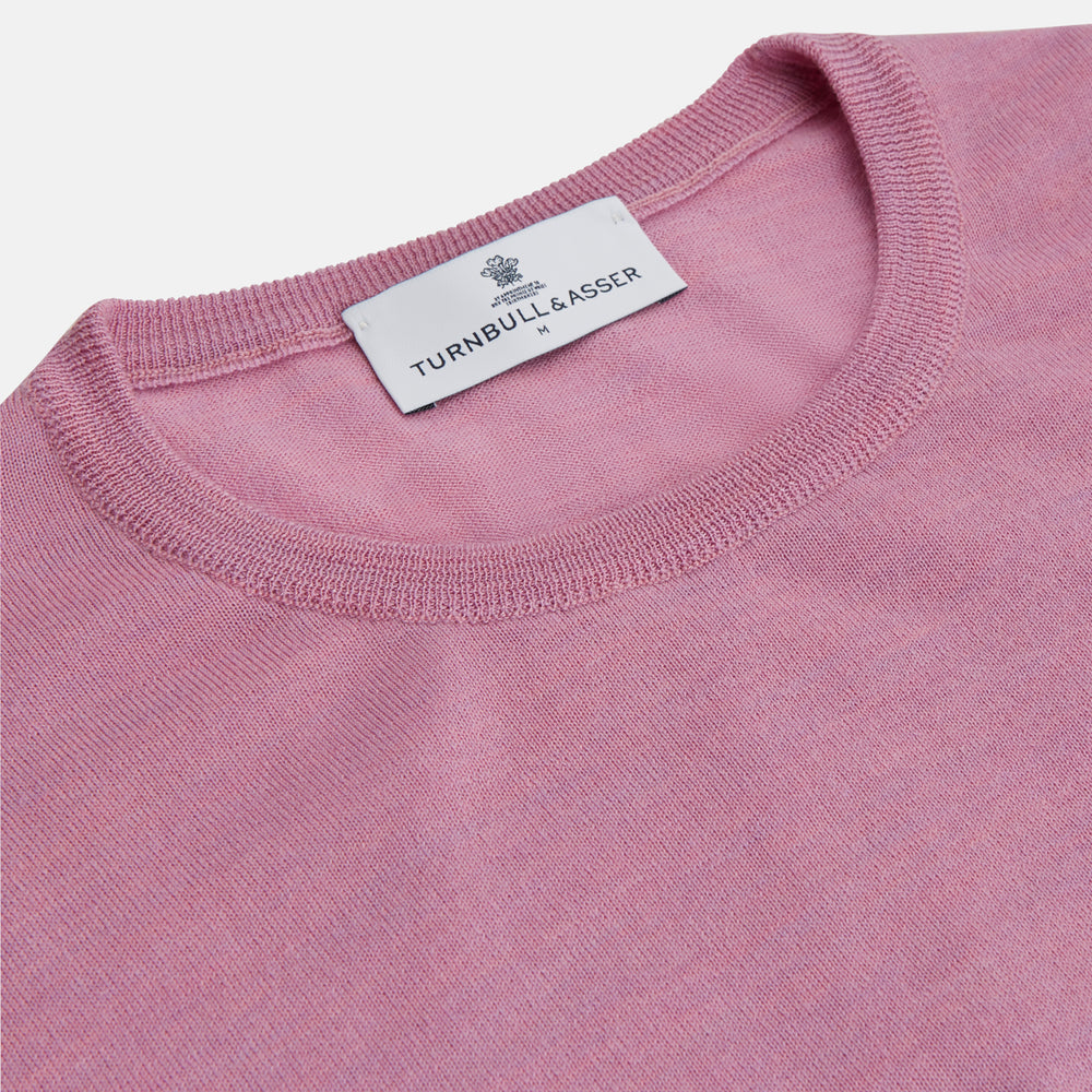 Wearing Turnbull & Asser’s Pink Merino Kingston Crew Neck Jumper, white pants, and a colorful scarf, a person stands against a plain light gray background with hands in their pockets.