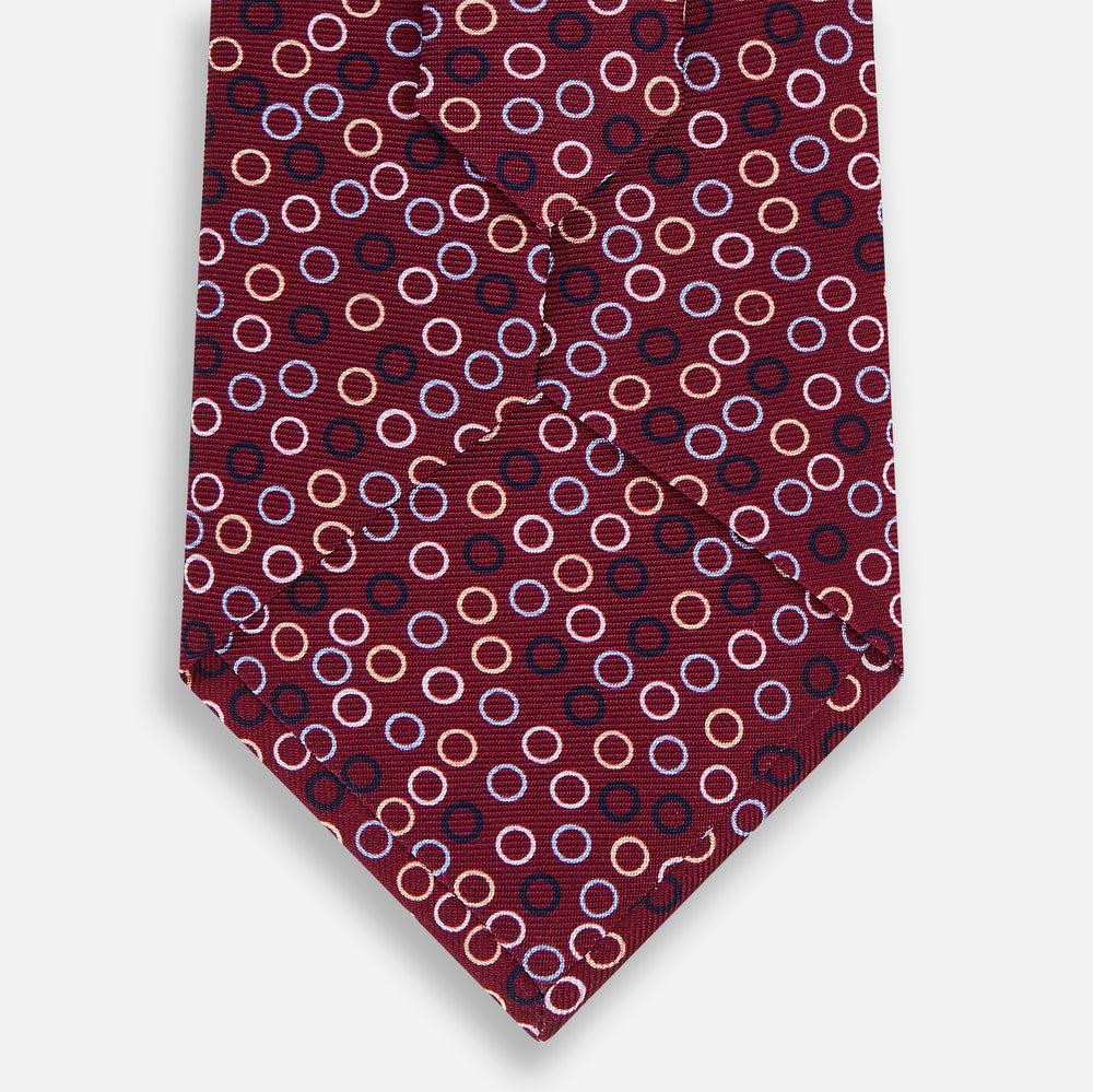 The Turnbull & Asser Pink and Navy Circle Burgundy Printed Silk Tie features overlapping circles in white, light blue, gold, and dark red, presented rolled up on a white background.