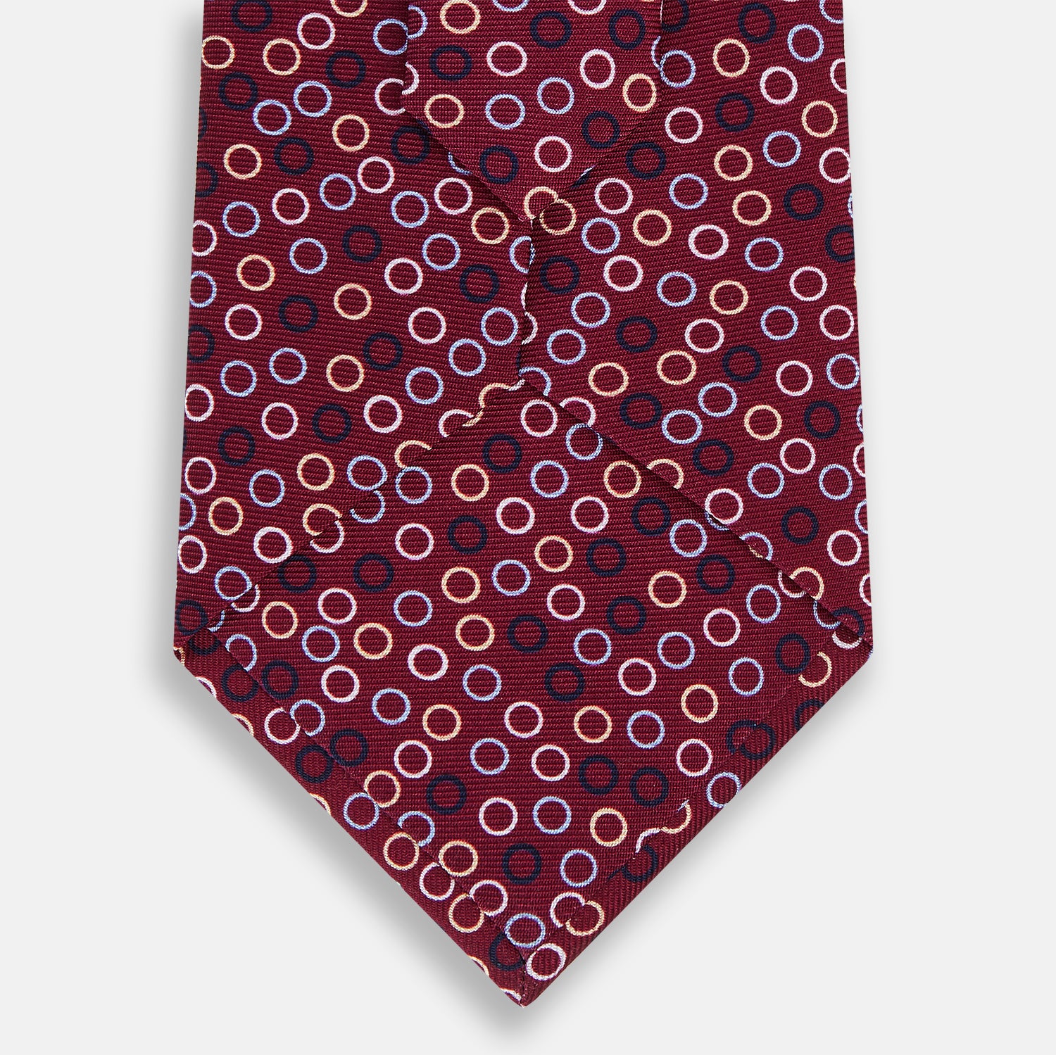 View 3 of product The Turnbull & Asser Pink and Navy Circle Burgundy Printed Silk Tie features overlapping circles in white, light blue, gold, and dark red, presented rolled up on a white background.