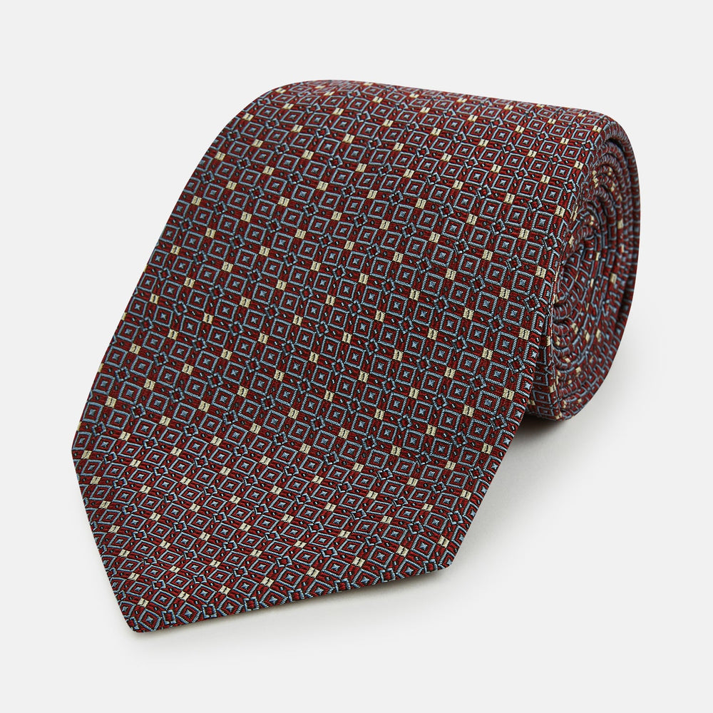 The Turnbull & Asser Burgundy Geometric Silk Tie, crafted in Britain, features a repeating pattern of light blue and white squares and diamonds. The neatly rolled tie is shown against a plain white background.