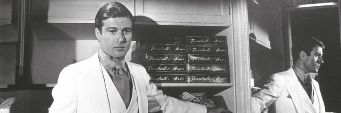 A man in a white suit stands in front of shelves stacked with boxes, looking at the camera, while his reflection appears in a mirror to his right. The scene is in black and white.