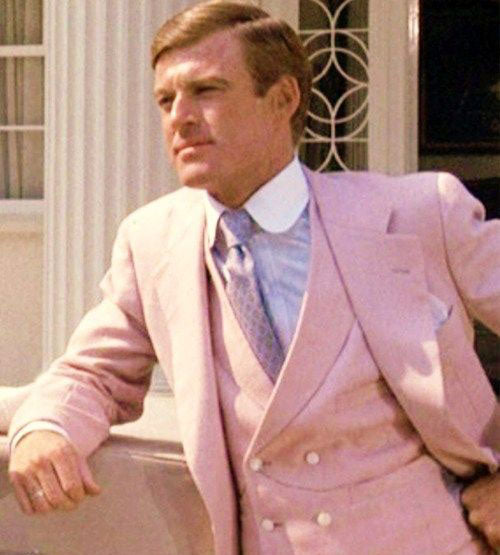 A man in a pale pink three-piece suit and tie leans against a white stone railing, looking to the side. White columns and decorative windows are visible in the background.