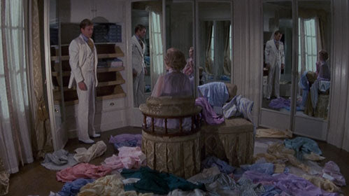 A man in a white suit stands by shelves, facing a woman seated at a vanity in a room with mirrored closets and clothing strewn across the floor. Both figures are reflected in the mirrors.