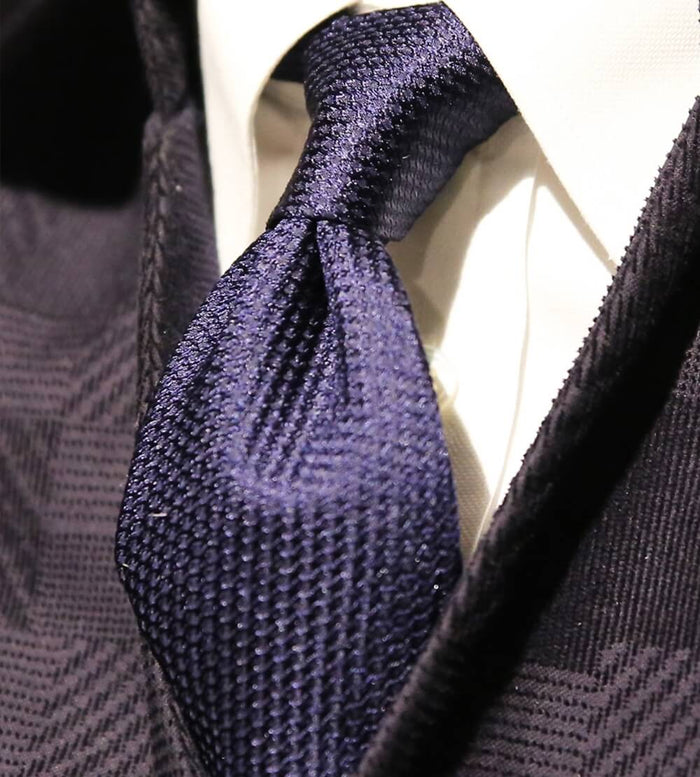 Close-up of a neatly tied textured navy blue tie on a white dress shirt, complemented by a dark patterned suit jacket.