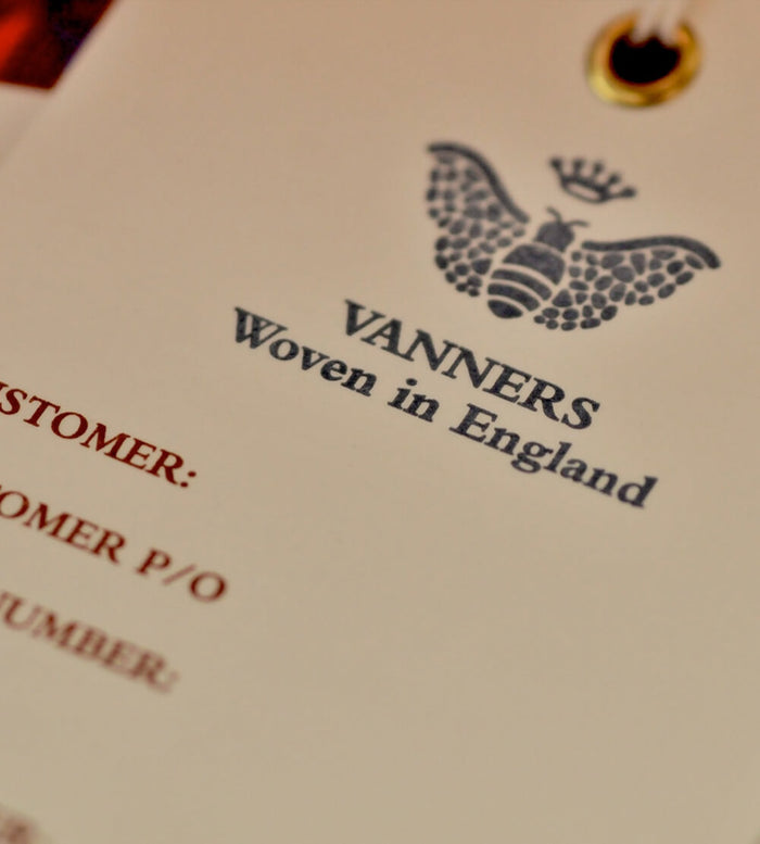 A clothing tag with the Vanners logo, featuring a bee and crown, and the text VANNERS Woven in England printed on it. Part of the tag includes red text for customer order details.