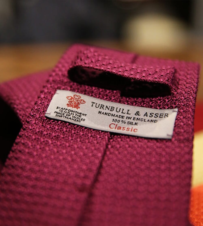 A close-up of a maroon textured silk tie with a label reading “Turnbull & Asser, Handmade in England, 100% Silk, Classic.”.
