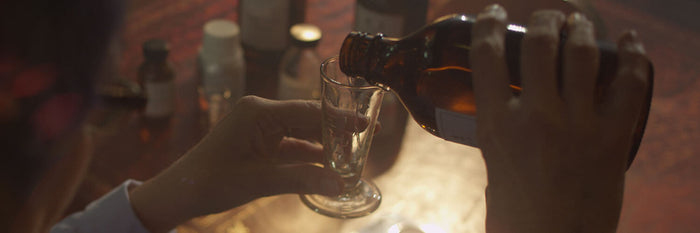 A person pours liquid from a brown glass bottle into a small clear glass on a wooden table with several other bottles in the background.