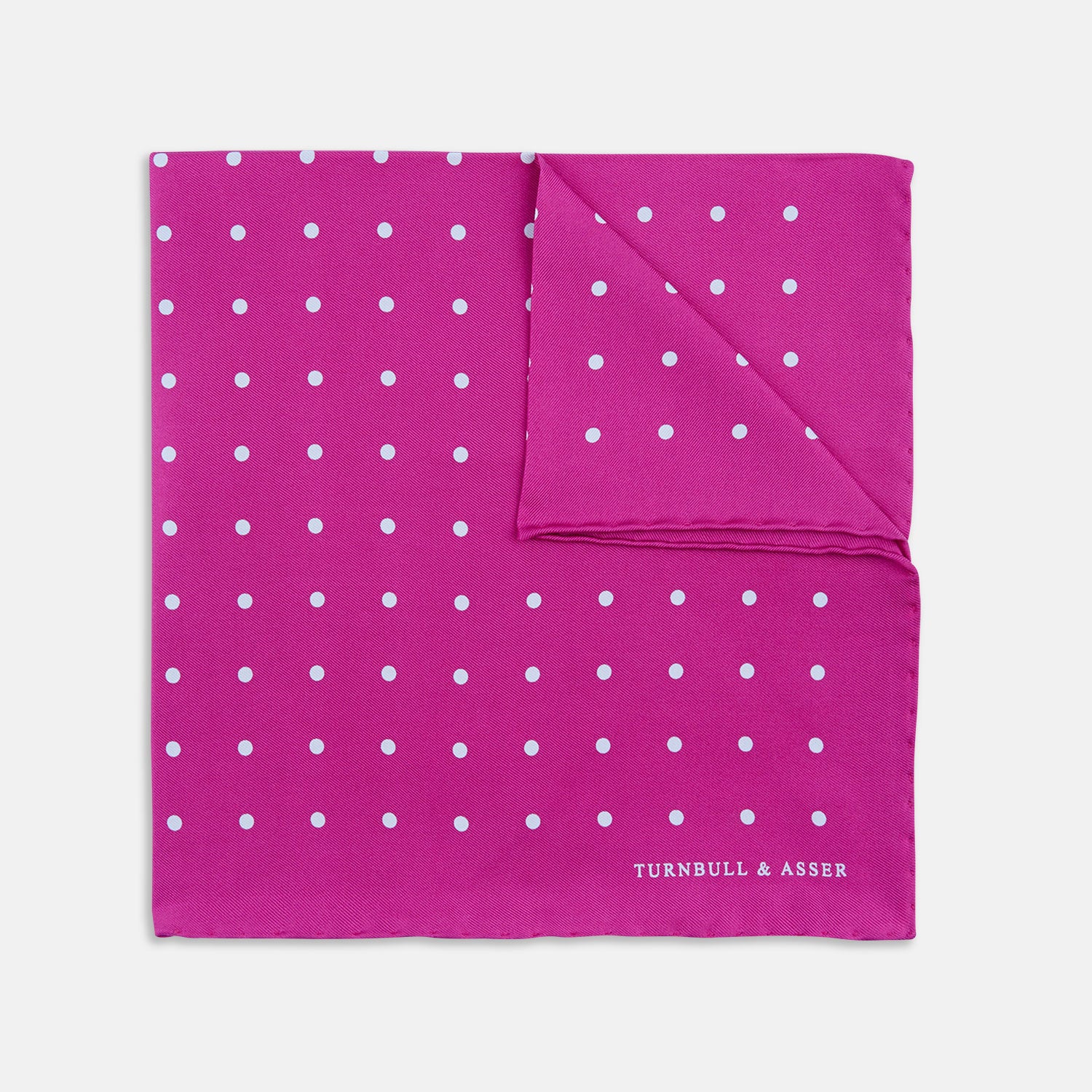 View 1 of product The Fuchsia Spot Silk Pocket Square by Turnbull & Asser features bright fuchsia silk with evenly spaced white spots and the brand’s name in one corner. A classic accessory, it is shown with one corner folded over.