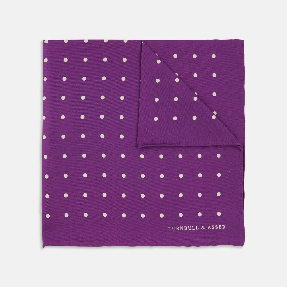 The Turnbull & Asser Purple Spot Silk Pocket Square features white polka dots and a small logo in the corner, with one corner folded over to showcase the underside.