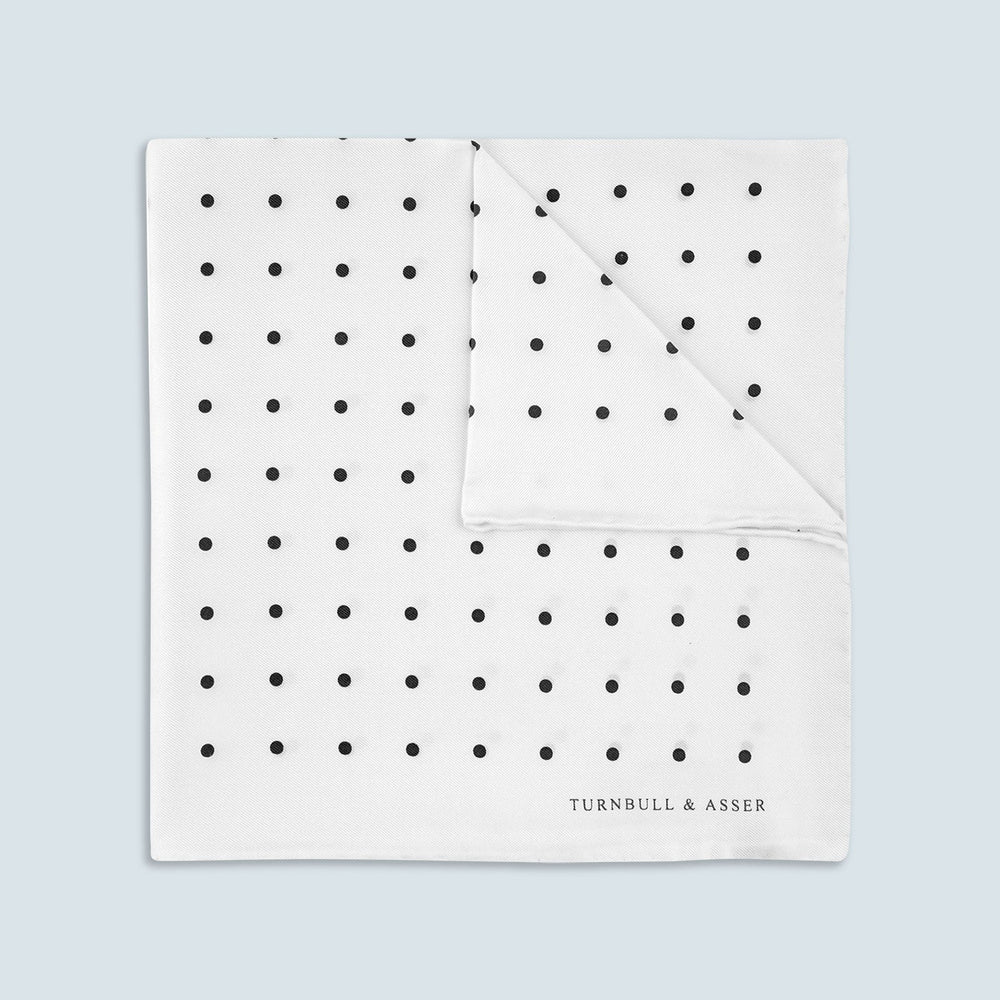 The Turnbull & Asser White and Black Spot Silk Pocket Square for men features evenly spaced black dots, a partial corner fold, and the brand name printed in black in the lower right corner.