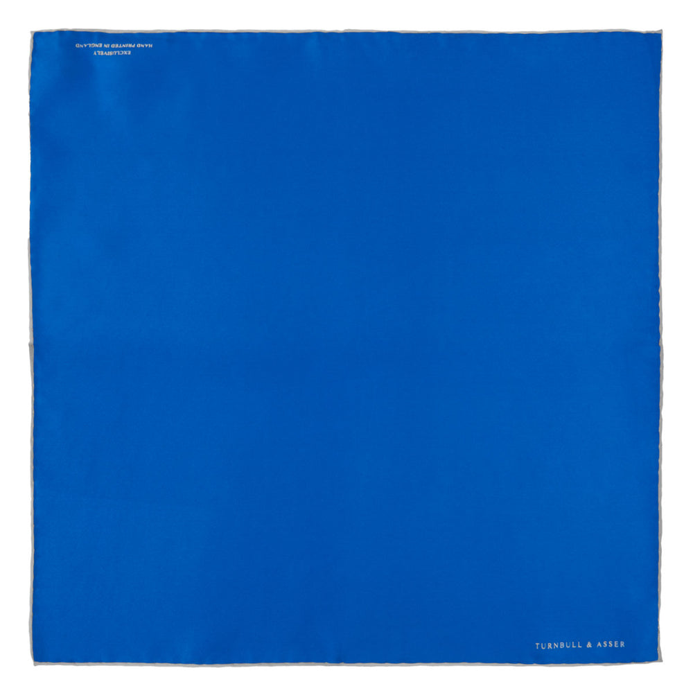The Royal Blue Piped Silk Pocket Square by Turnbull & Asser features a vibrant blue hue, subtle stitched border, and the brand name printed in white in the lower right corner.