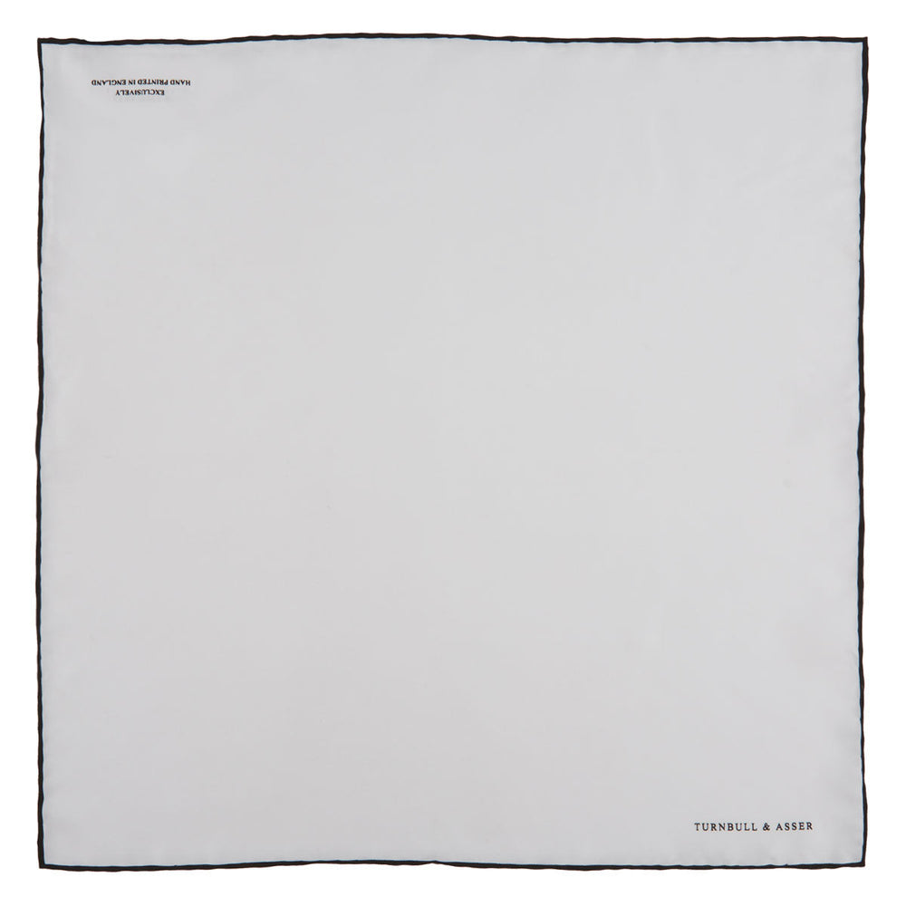The Turnbull & Asser White and Black Piped Silk Pocket Square features a crisp white base with black edging, neatly folded to showcase the logo in black on the lower right—elegant monochrome style for any occasion, shown on a light gray background.