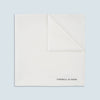 View 1 of product The White Piped Silk Pocket Square by Turnbull & Asser is shown folded at one corner with the brand's logo in black on the lower right, set against a light blue background.
