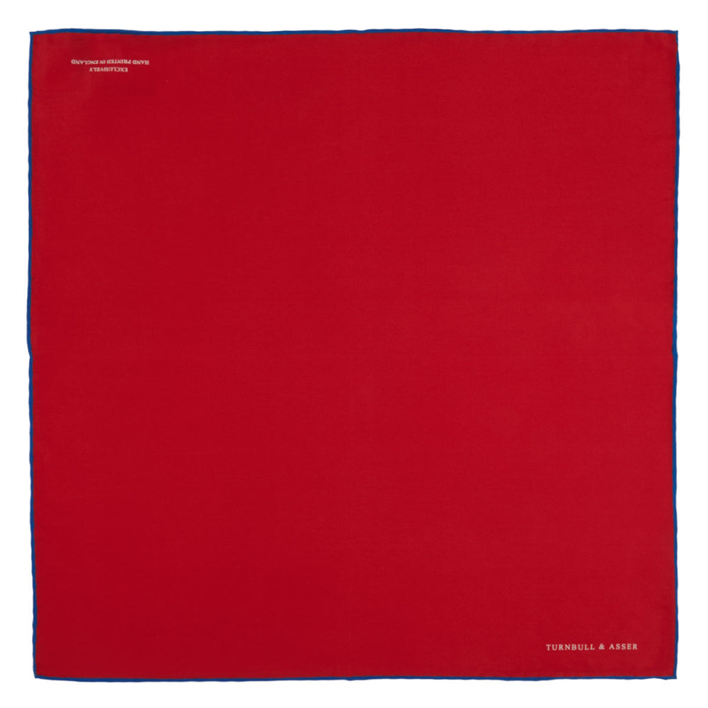 The Red and Navy Piped Silk Pocket Square by Turnbull & Asser features classic red silk with navy edging and the brand’s name printed in white on the bottom right corner.