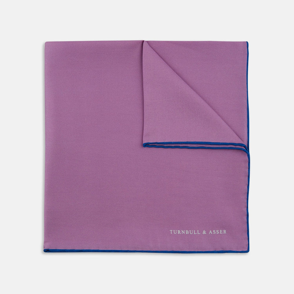 The Lilac and Navy Piped Silk Pocket Square by Turnbull & Asser features a folded lilac design with dark blue trim and the brand's name printed in white in the lower right corner.