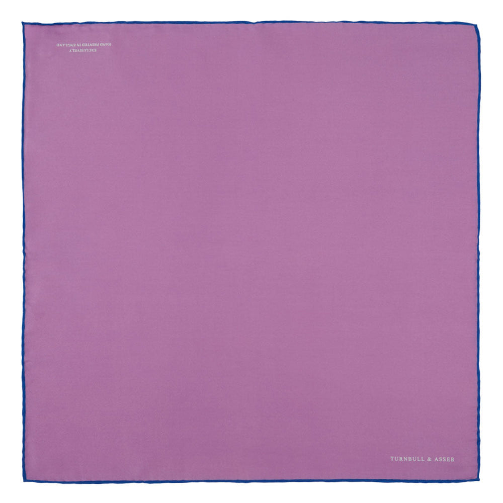 The Lilac and Navy Piped Silk Pocket Square by Turnbull & Asser features a folded lilac design with dark blue trim and the brand's name printed in white in the lower right corner.