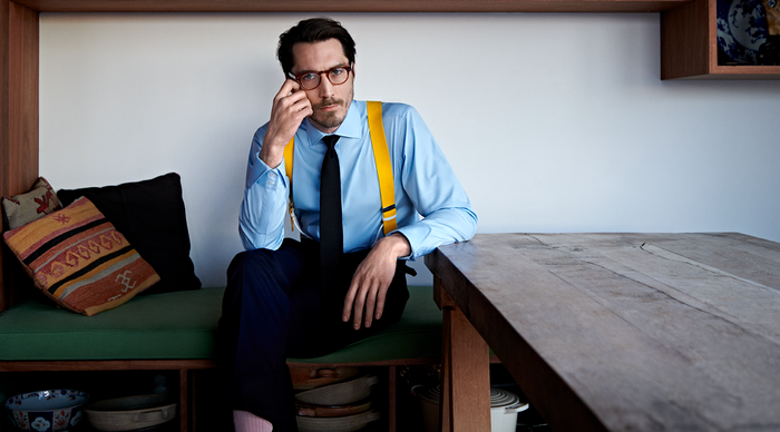 A man in a blue shirt, yellow suspenders, and a black tie sits on a bench by a wooden table, holding his glasses and looking at the camera. Pillows and shelves with bowls are in the background.