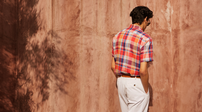 A man wearing a red, blue, and white plaid short-sleeve shirt and white pants stands with his back to the camera, hands in pockets, next to a textured, sunlit terracotta wall.
