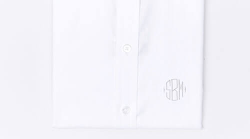 A folded white dress shirt with two visible buttons and a round, gray monogram SRM embroidered near the bottom right side.