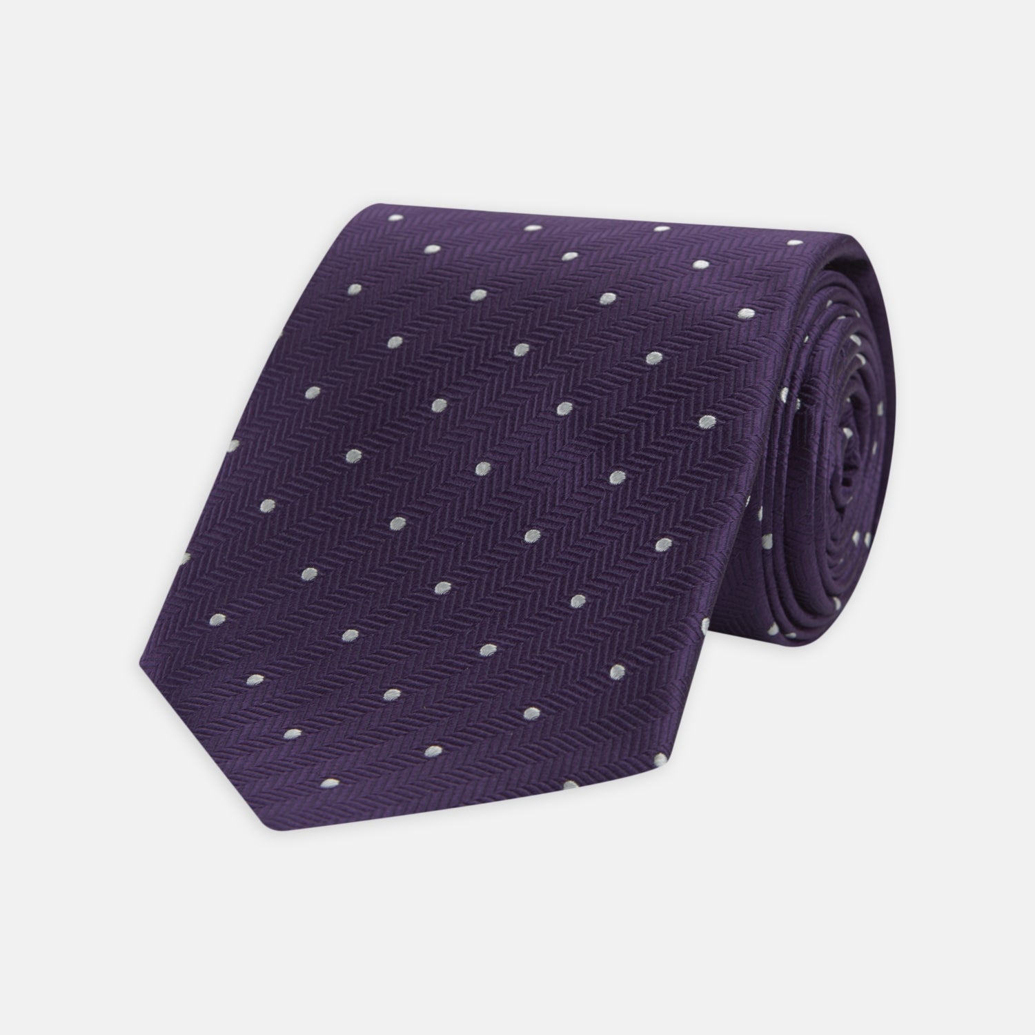 View 1 of product The Turnbull & Asser Plum and White Small Spot Herringbone Silk Tie is shown rolled up on a plain white background, featuring a classic silk herringbone weave in plum with small white spots.