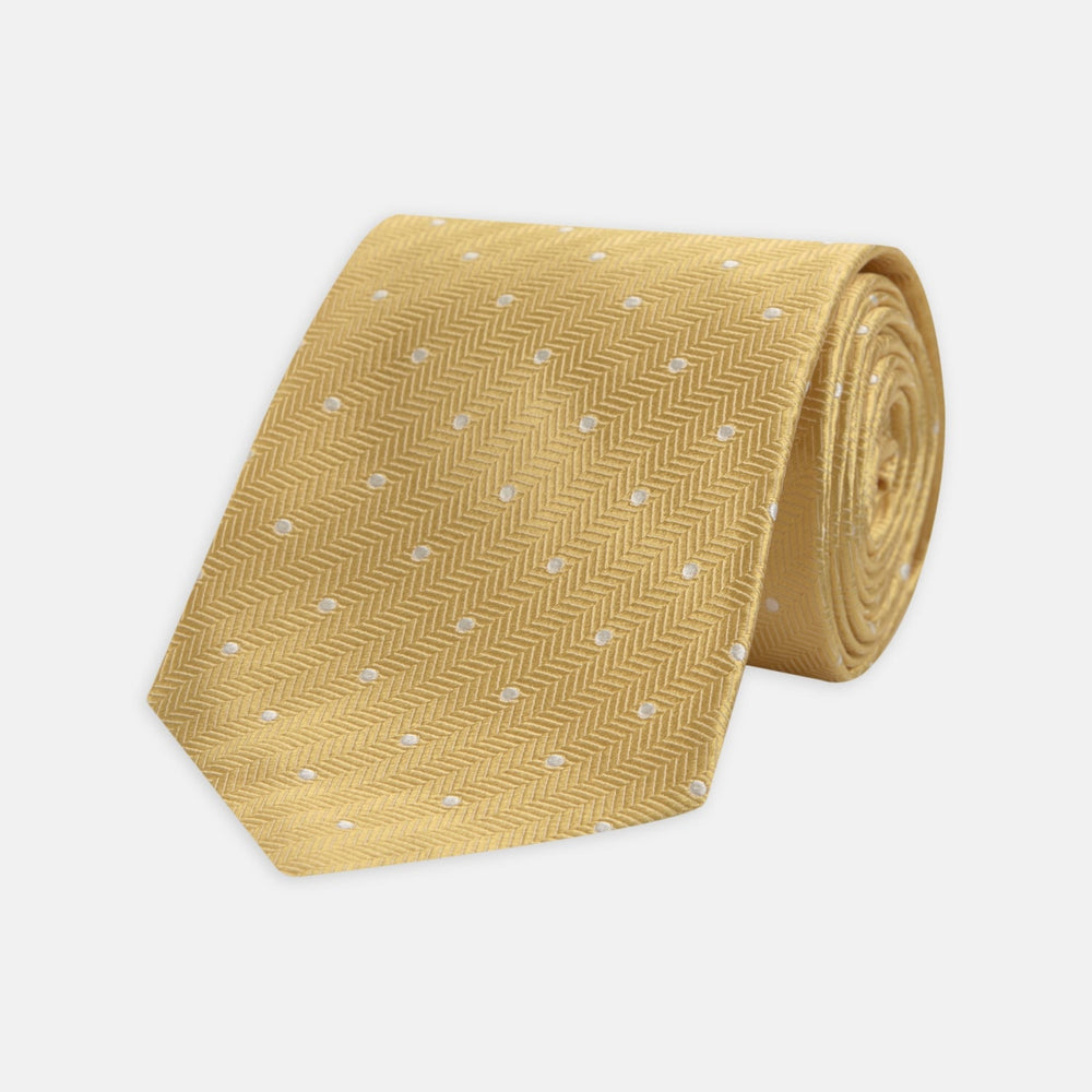 The Turnbull & Asser Gold and White Small Spot Herringbone Silk Tie is crafted from fine English silk, featuring a gold herringbone weave with white polka dots, and is shown rolled up on a white background.