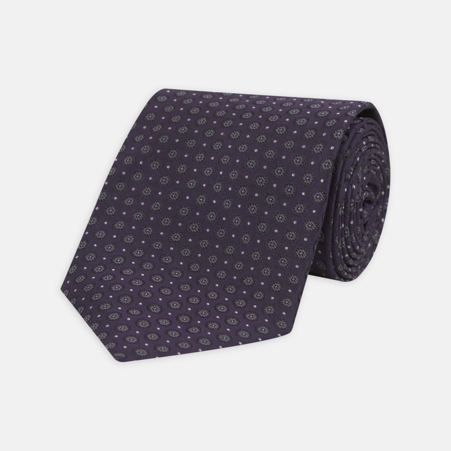 View 1 of product The Turnbull & Asser Plum Floral Spot Silk Tie features a rolled-up classic design with small, evenly spaced floral spots in lighter purple and white, ideal for office shirts and shown against a plain white background.