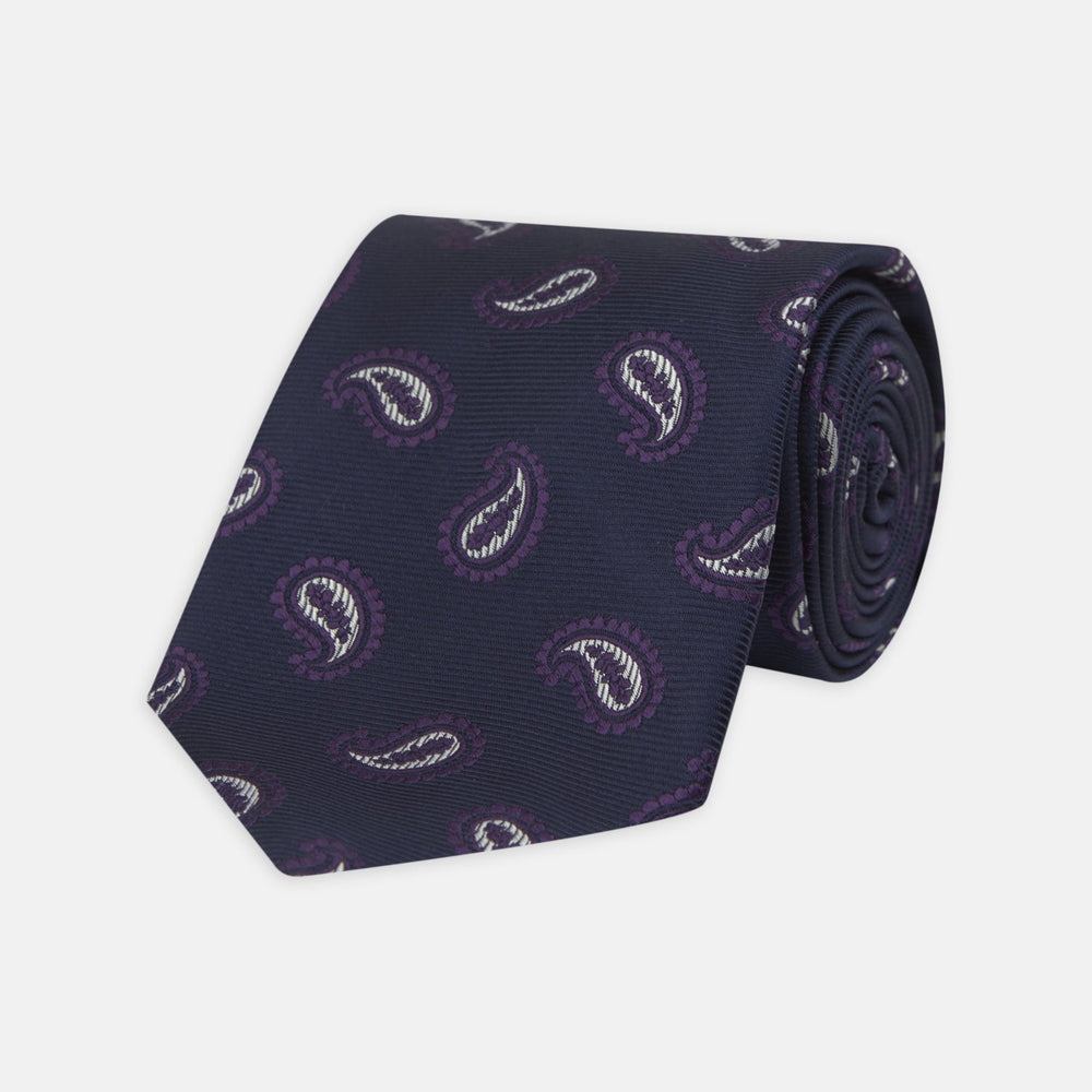 The Turnbull & Asser Navy and Plum Floating Paisley Silk Tie features a repeating purple and white paisley pattern, elegantly rolled up on a white background.