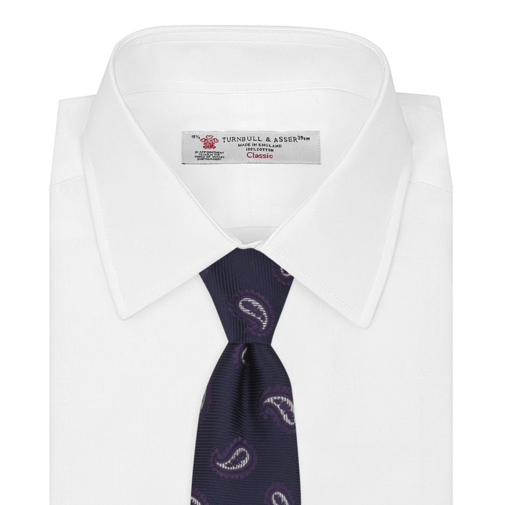 The Turnbull & Asser Navy and Plum Floating Paisley Silk Tie features a repeating purple and white paisley pattern, elegantly rolled up on a white background.