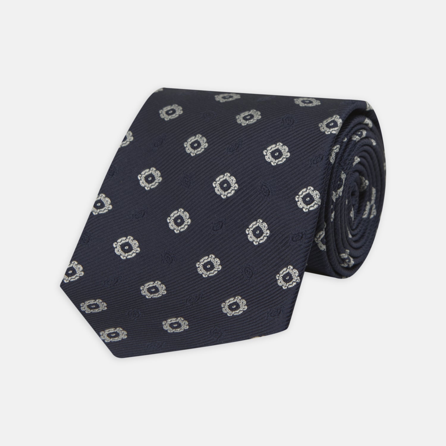 View 1 of product The Turnbull & Asser Navy and Silver Emblem Spot Silk Tie features a refined repeating emblem pattern in silver on navy silk, making it an elegant accessory for any office shirt.