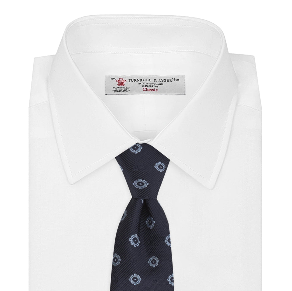 The Turnbull & Asser Navy and Light Blue Emblem Spot Silk Tie features a classic rolled design with a repeating light blue emblem pattern, ideal for office wear, shown against a plain white background.