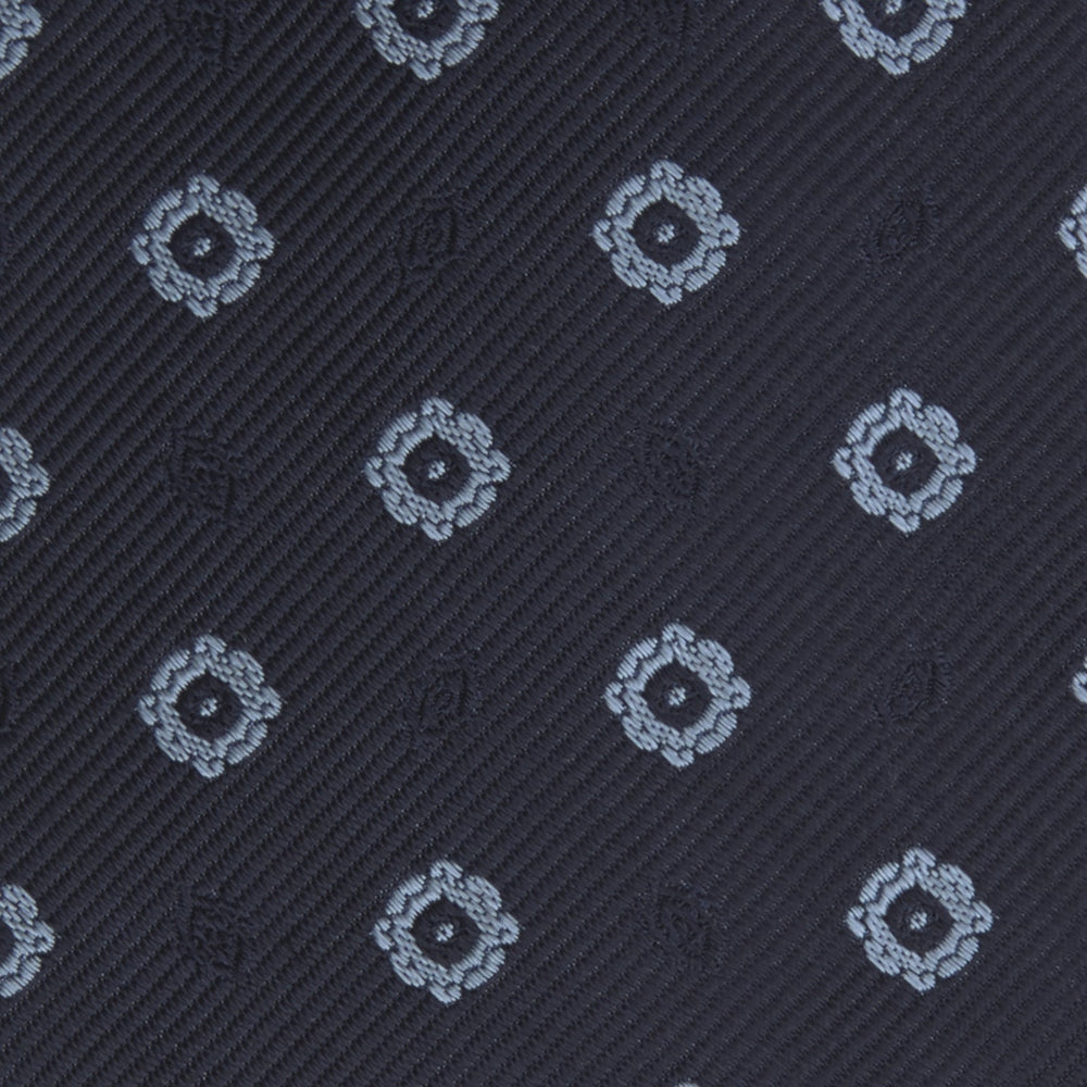 The Turnbull & Asser Navy and Light Blue Emblem Spot Silk Tie features a classic rolled design with a repeating light blue emblem pattern, ideal for office wear, shown against a plain white background.