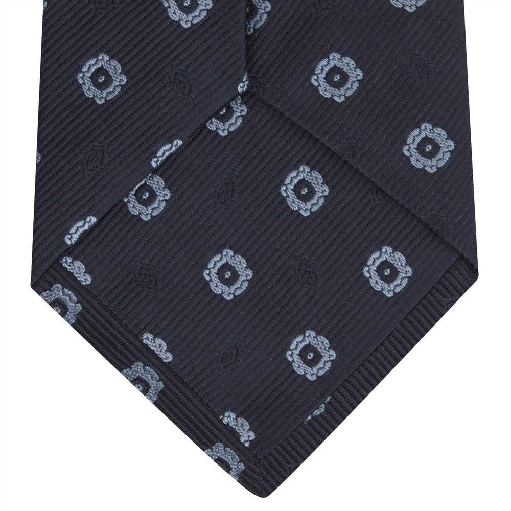 The Turnbull & Asser Navy and Light Blue Emblem Spot Silk Tie features a classic rolled design with a repeating light blue emblem pattern, ideal for office wear, shown against a plain white background.
