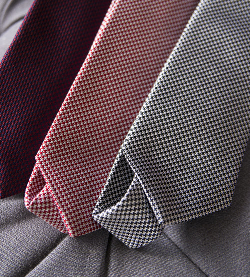 Three textured neckties in shades of dark red, maroon, and black-and-white houndstooth are laid out diagonally on a light gray suit jacket.