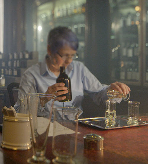 A person with short gray hair and glasses pours liquid from a bottle into a glass at a laboratory table, surrounded by glassware and scientific equipment. Shelves with bottles are visible in the background.