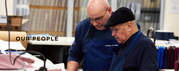 Two older men wearing glasses and blue aprons work together in a fabric workshop. The text “OUR PEOPLE” appears on the left. Various textiles and tools are visible in the background.